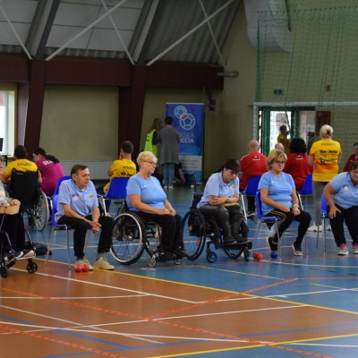 Zdjęcie przedstawia ogólny rzut na salę sportową gdzie osoby siedzące na wózkach inwalidzkich rozgrywają zawody Boccia